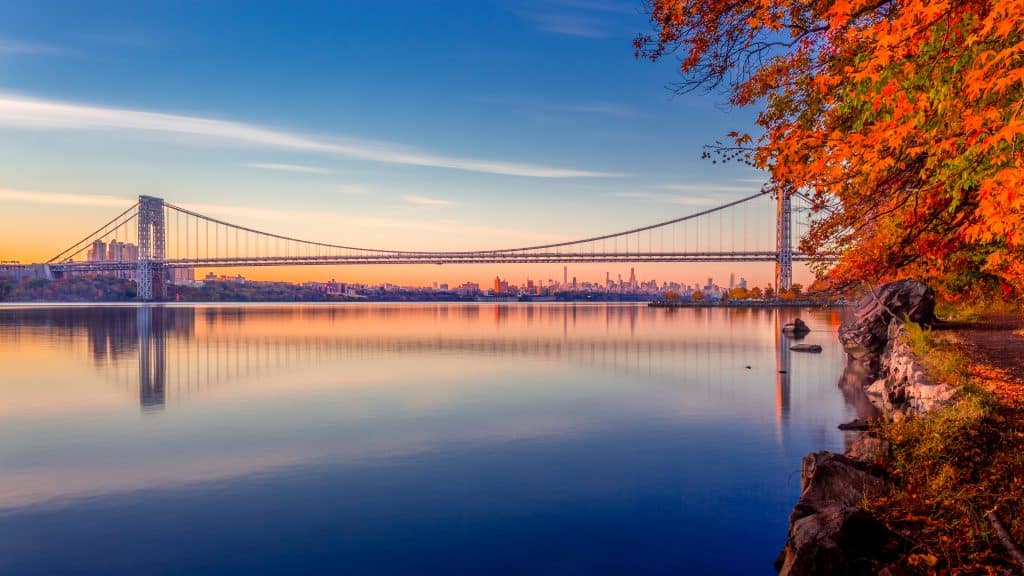 Autumn foliage along a calm river with a suspension bridge and a city skyline in the distance during sunset. one of the most famous bridges in New York City. 