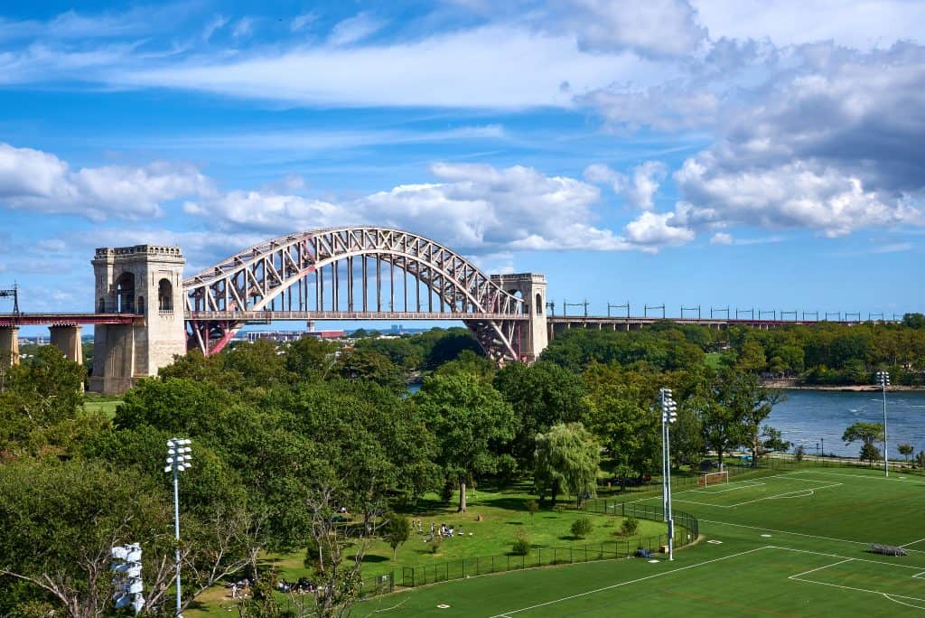 A steel arch bridge spanning over a river with adjacent park and sports field on a sunny day with clouds. Famous bridges in New York.