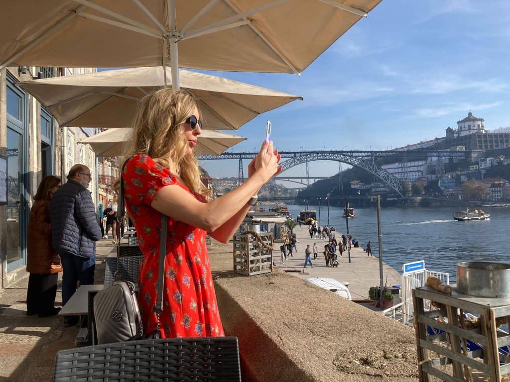 Woman in a red dress taking a photo with her smartphone on a riverside terrace, with a bridge and cityscape in the background.