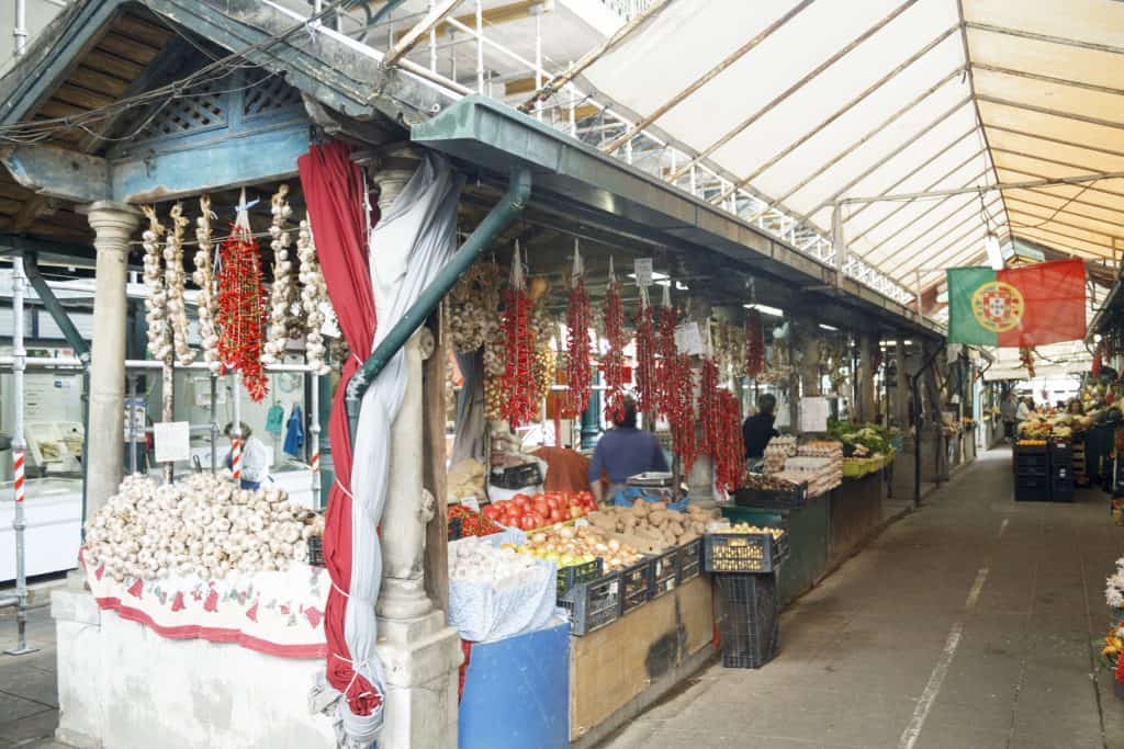 An outdoor market stall displaying an array of garlic and red chili pepper strings under a metal roof structure.