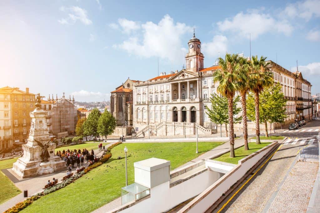 Sunny view of a historic city square with an ornate classical building, a statue, palm trees, and people walking. sun flares visible.