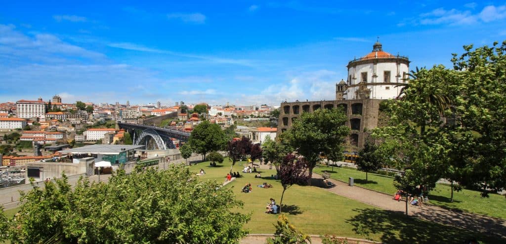 View of a busy urban park in porto, with an old church, modern bridge, and downtown skyline under a clear blue sky.