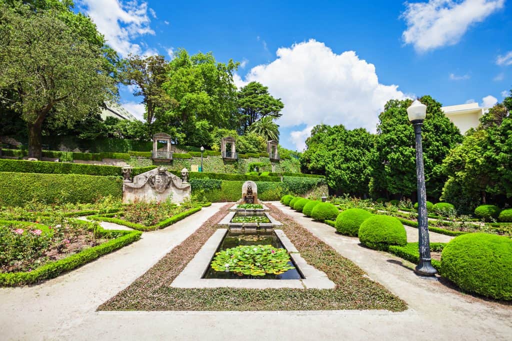 Formal garden with symmetrical design, featuring trimmed hedges, a central fountain with water lilies, and a cloud-filled blue sky.