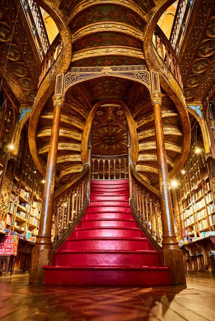 Ornate wooden staircase with red carpet in a richly decorated library, featuring elaborate ceiling paintings and book-lined walls.