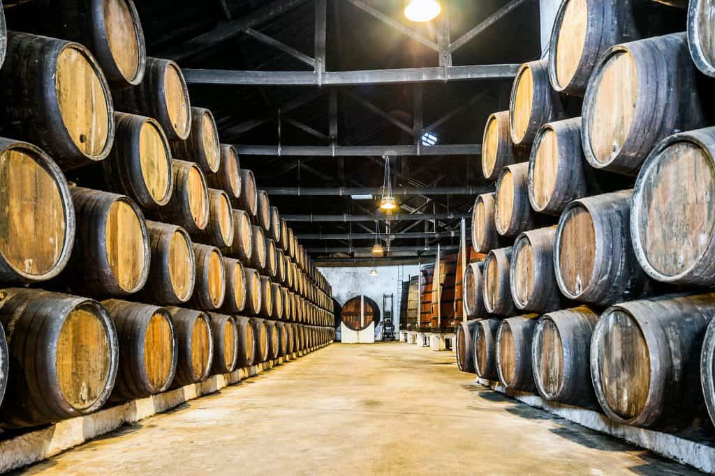 Rows of wooden barrels stacked in a dimly lit distillery warehouse with a central walkway leading to an arched door. Vila Nova de Gaia in Porto.