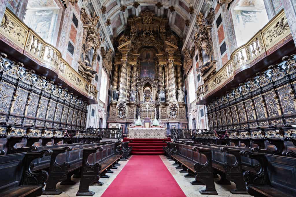 Interior of a baroque church with elaborate golden altar, red carpet aisle, and ornately carved wooden pews.