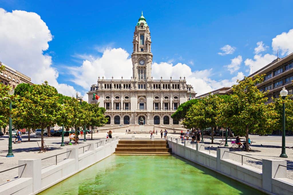 View of the porto city hall in portugal, with its distinctive clock tower, surrounded by trees and a water feature in the foreground, on a sunny day with people around.