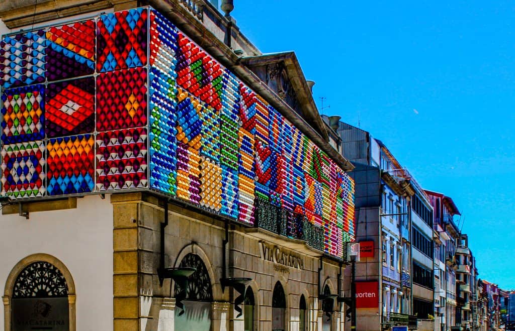 Colorful mosaic facade on a building in an urban street lined with shops and architectural detailing under a clear blue sky. Street art in Porto.