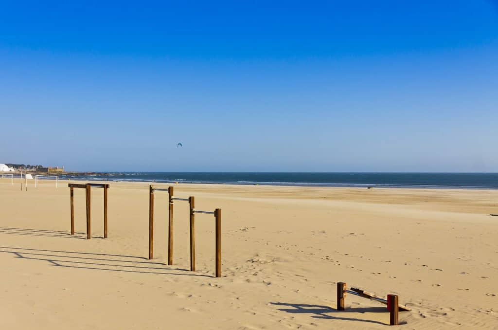 A serene beach scene with a clear blue sky, sandy shore, and calm sea. metal structures partially buried in sand are visible, and a single bird flies in the distance.