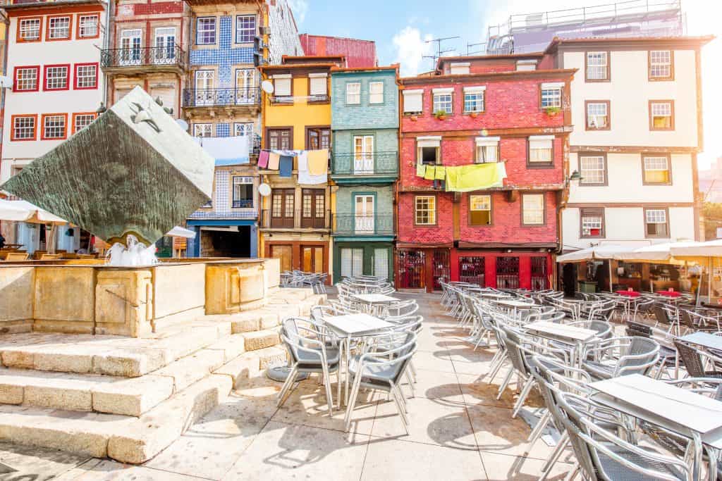 Colorful buildings and café terrace with many empty chairs in a sunny european square, stone pavement and large modern sculpture dominating the foreground. Ribeira square Porto.