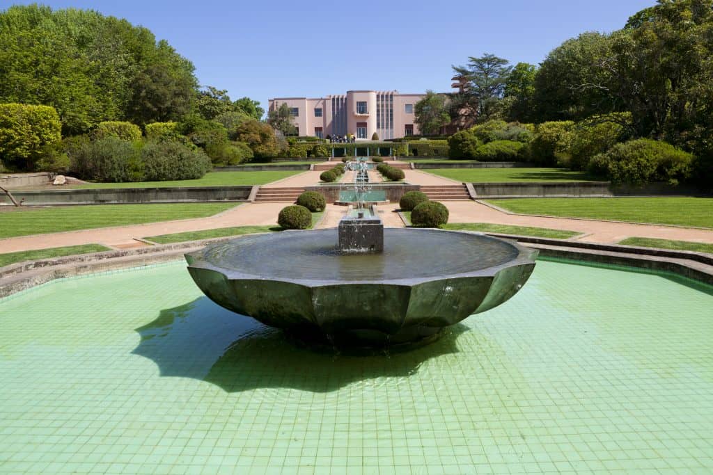Symmetrical view of a formal garden featuring a large central fountain, leading to a pink-hued building in the background, surrounded by lush greenery. Serralves Foundation in Porto.