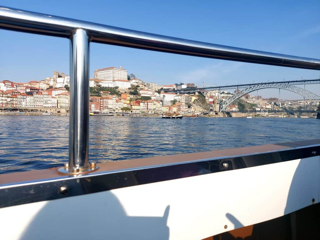 View of porto from a boat, featuring the cityscape and dom luis i bridge, with clear skies and a stainless steel railing in the foreground.