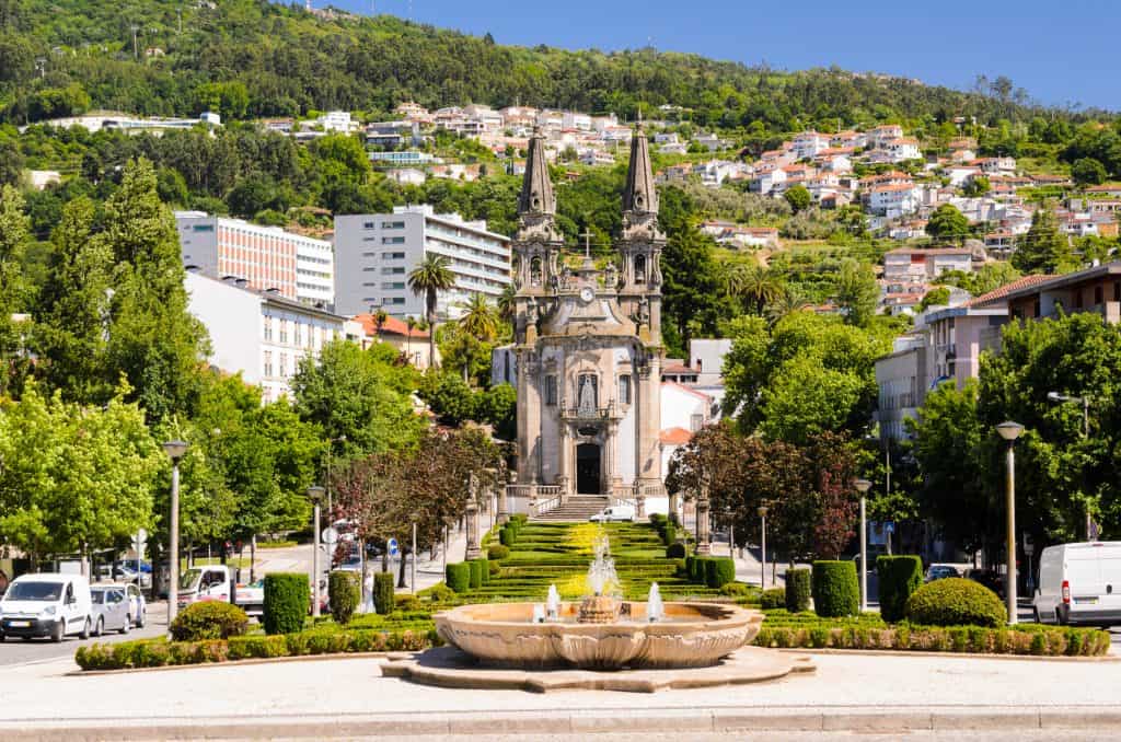 A view of the gothic-style lamego fountain in front of a tree-lined staircase leading to a hill, with houses scattered across the hillside under a clear blue sky.