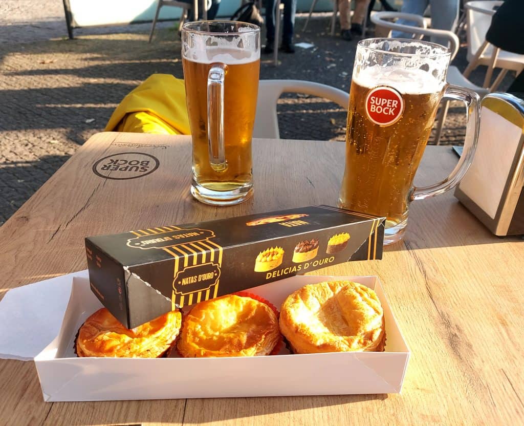 Three glasses of beer and a box of portuguese custard tarts on a wooden table at an outdoor cafe.