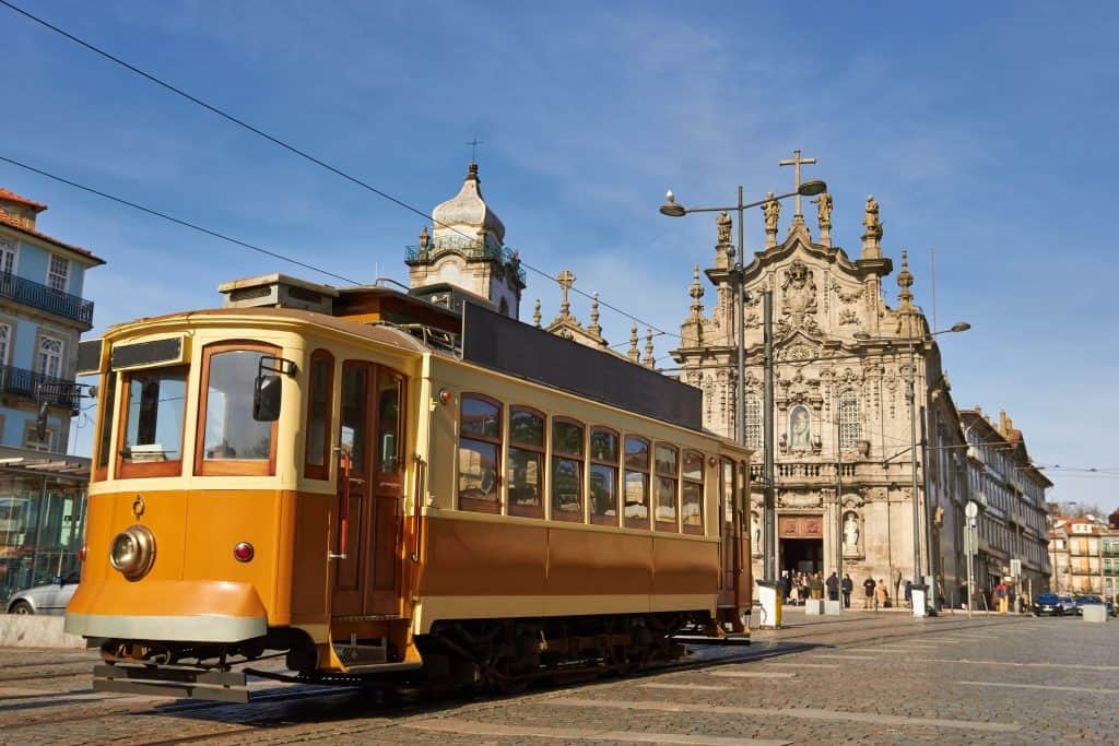 A vintage tram rolling in front of a baroque church under a clear blue sky in a cobblestoned square.
