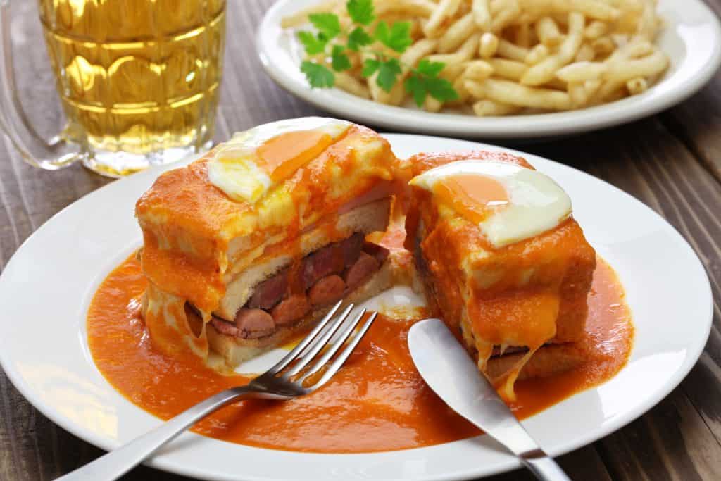 A plate containing a slice of francesinha, a portuguese sandwich with various meats, cheese, and an egg, served with beer and fries.