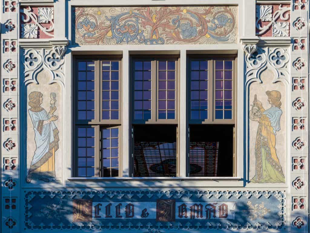 Ornate building facade with three stained glass windows framed by intricate murals and decorative tiles featuring artwork and text "fides" and "spes.