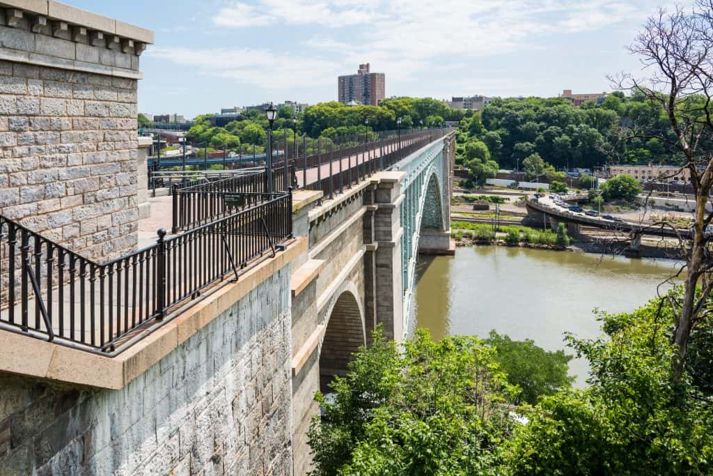 View of a stone arch bridge spanning the Harlem river in NYC, with lush greenery on the banks and a cityscape in the background.