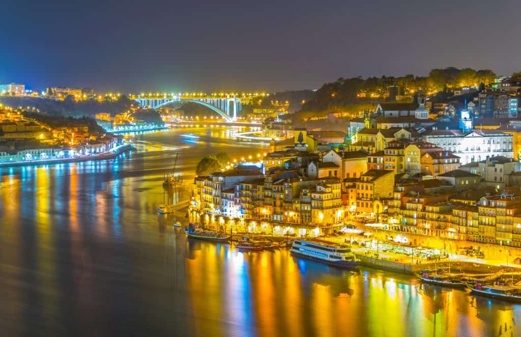 Nighttime view of the Porto cityscape by a river, featuring illuminated buildings and bridges, and boats docked along the waterfront.
