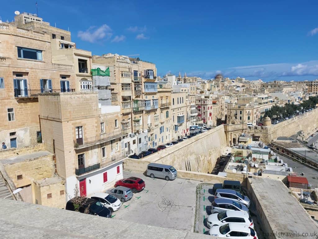 View of a steep, sunlit street lined with traditional limestone buildings in Valletta, Malta, under a clear blue sky.