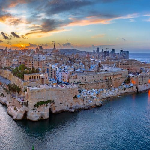 Experience a panoramic view of Valletta at sunset, featuring historic buildings on a peninsula embraced by calm waters, with the city skyline as a breathtaking backdrop. Discover things to do in Valletta while soaking in its rich history and stunning vistas.