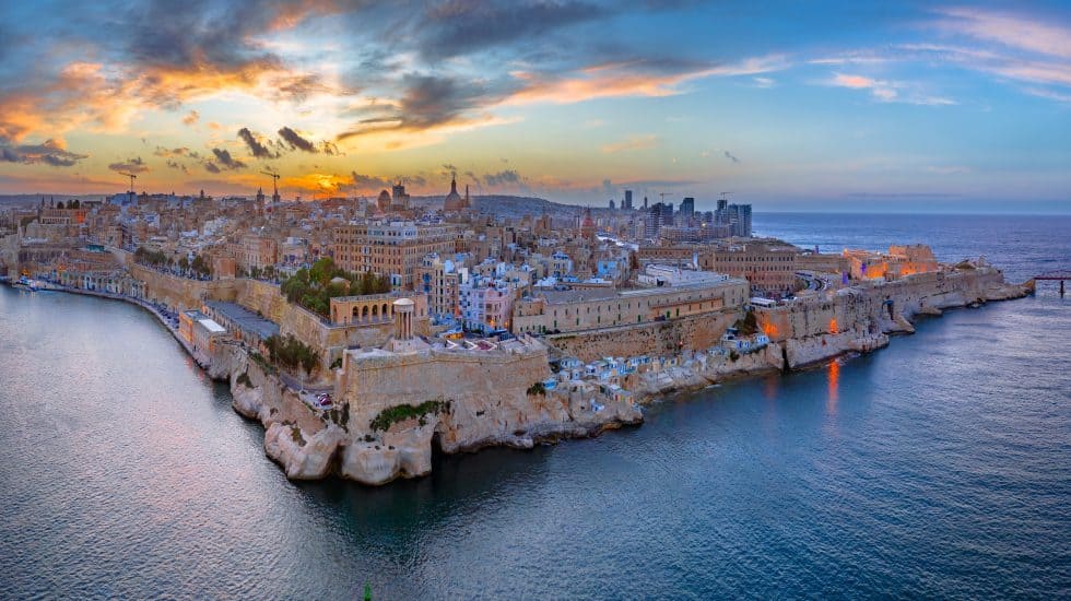 Experience a panoramic view of Valletta at sunset, featuring historic buildings on a peninsula embraced by calm waters, with the city skyline as a breathtaking backdrop. Discover things to do in Valletta while soaking in its rich history and stunning vistas.