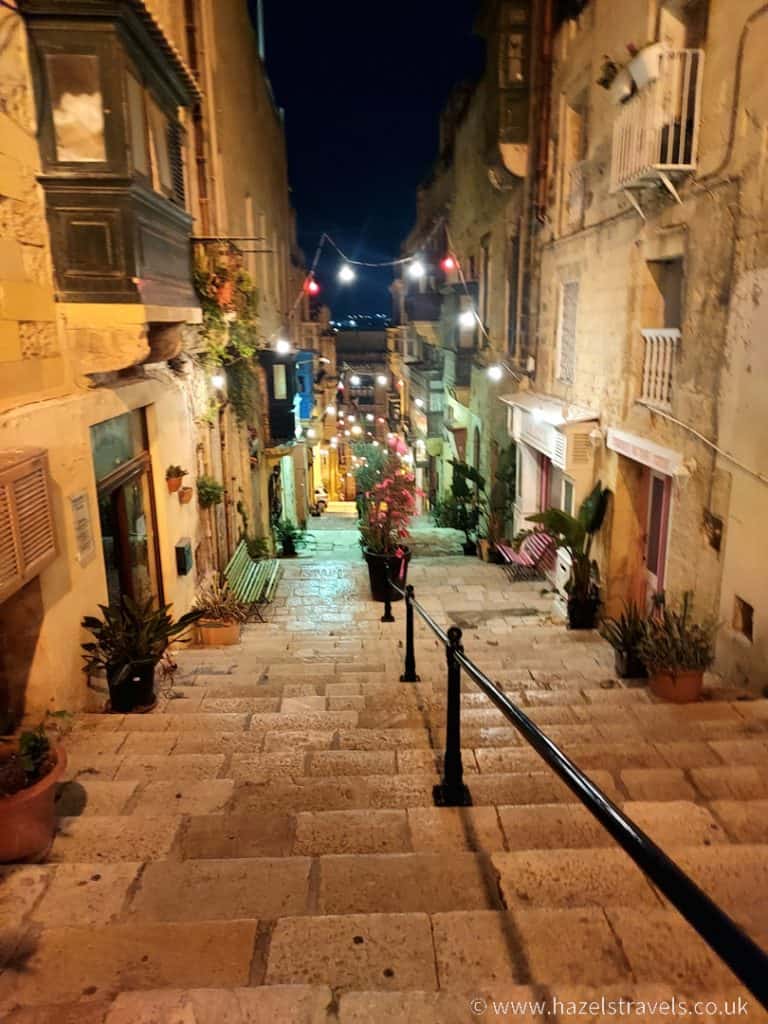 A nighttime view of a narrow, steep cobblestone street lined with old buildings and decorated with small lights, seen in Valletta, Malta.