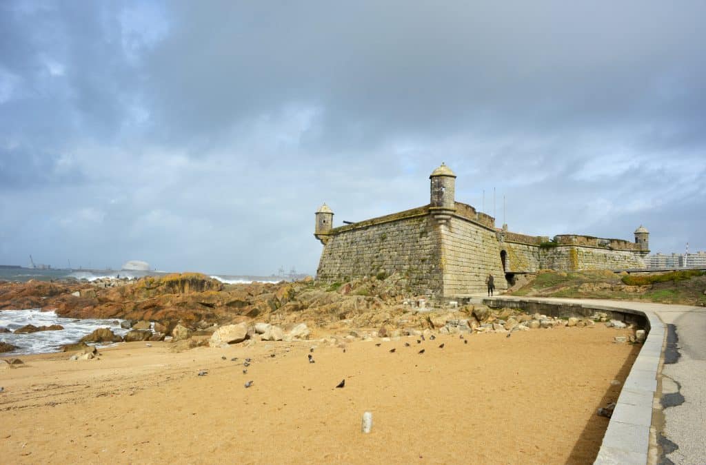 Stone fortress on a sandy beach in Porto with rough sea waves, under a cloudy sky. a winding path leads up to the fort.