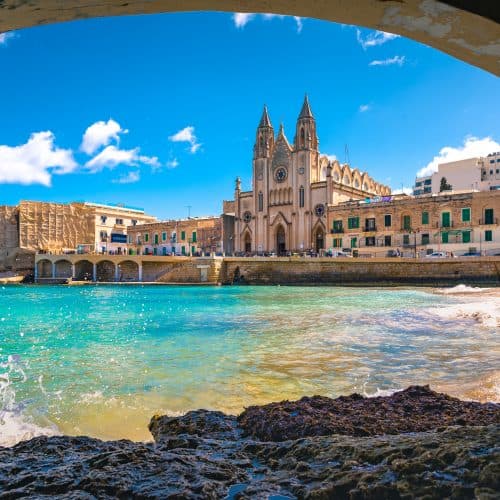 View of a beachside area in Malta with clear turquoise water and historic buildings, including a prominent cathedral, under a bright blue sky.