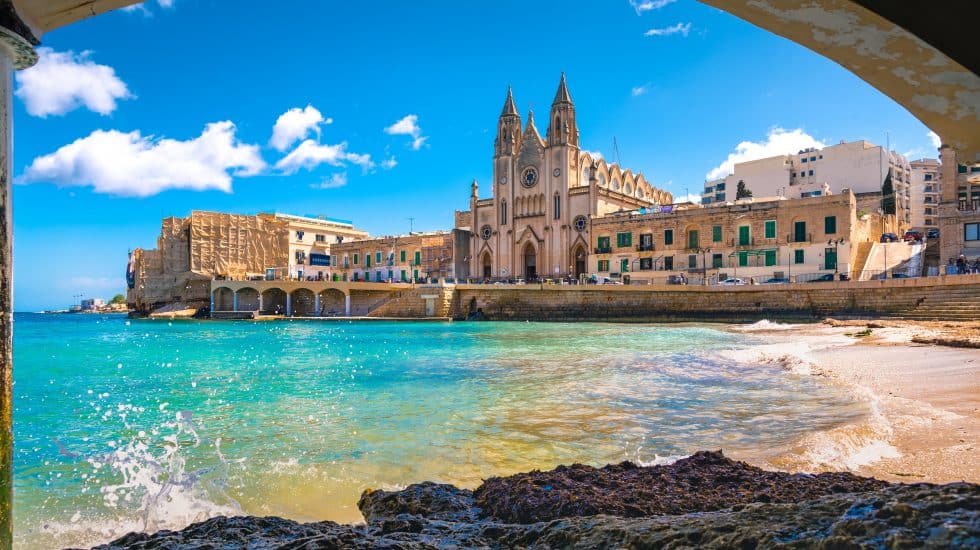 View of a beachside area in Malta with clear turquoise water and historic buildings, including a prominent cathedral, under a bright blue sky.