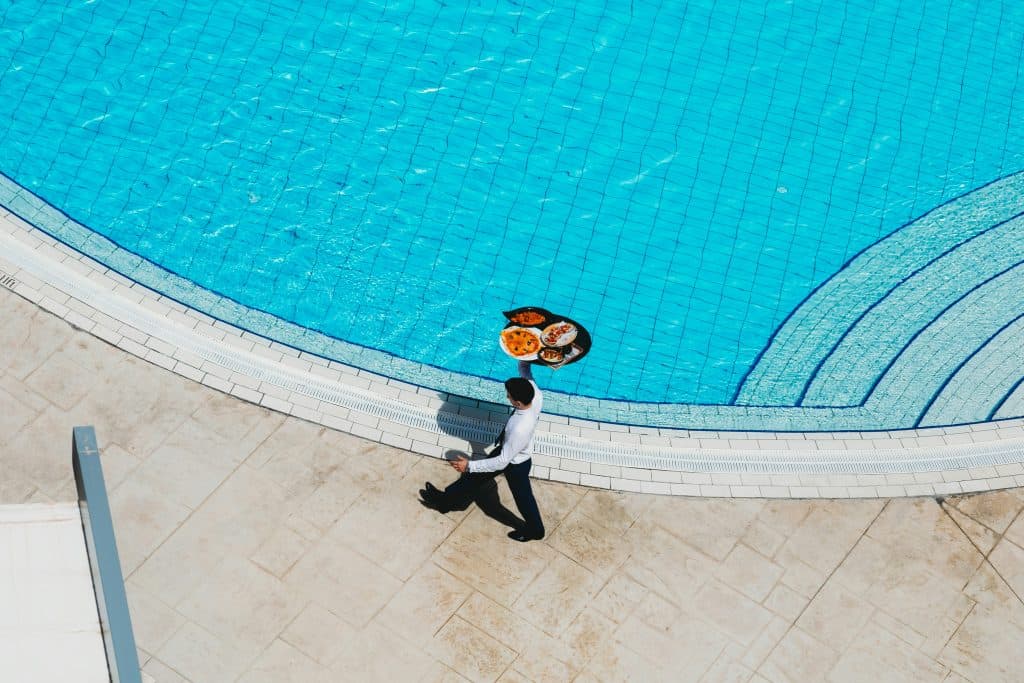 A person carries a tray with food and drinks by a blue swimming pool at the Hilton Malta, walking on a beige tiled surface under a clear sky. 