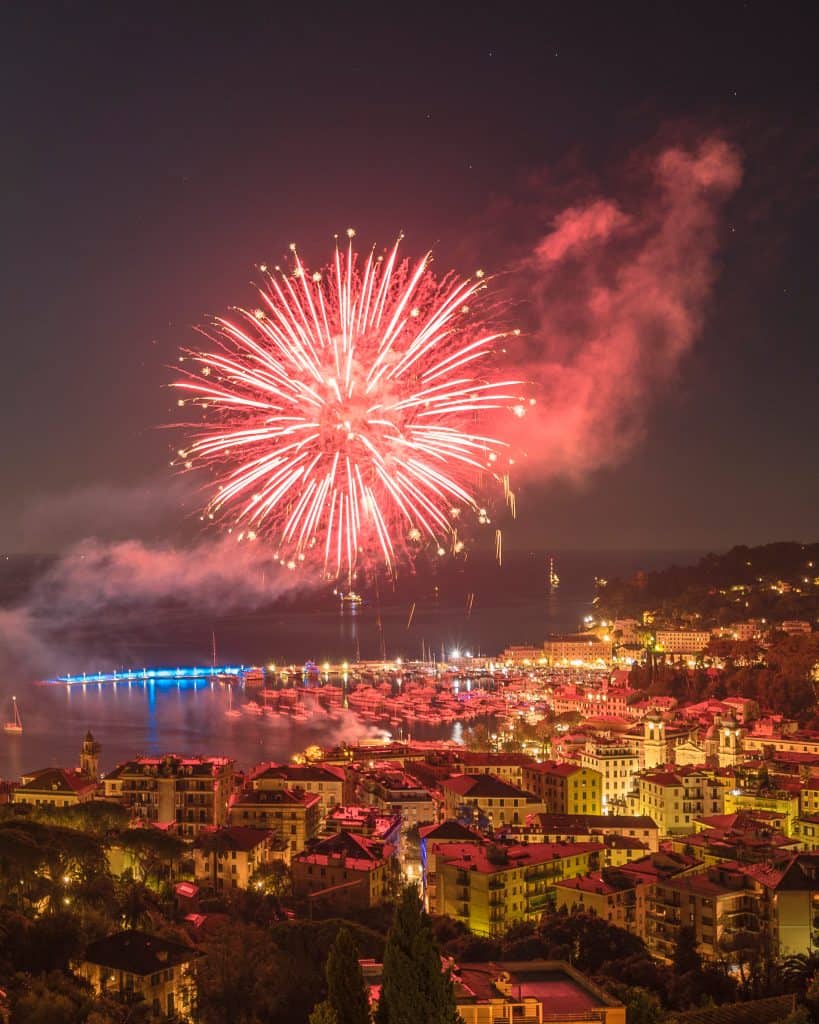 A bright red firework display illuminates the night sky over a coastal town, with lights from buildings and boats reflecting on the water below.