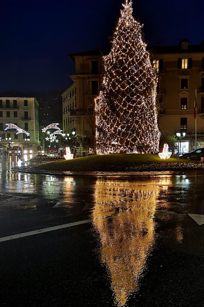 A large, brightly lit Christmas tree in a town square at night, with light decorations on nearby buildings and a reflection of the tree on the wet pavement.