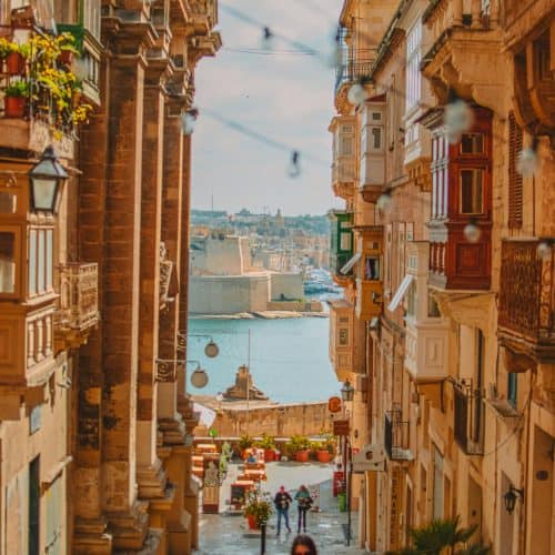 A narrow street in Valletta lined with buildings and balconies leads down to a waterfront with a body of water and buildings in the background under a clear sky.