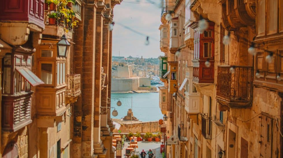 A narrow street in Valletta lined with buildings and balconies leads down to a waterfront with a body of water and buildings in the background under a clear sky.