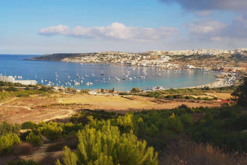 A coastal town (Mellieħa in Malta) with numerous boats anchored in a calm bay, surrounded by rolling hills, greenery, and some clouds in the blue sky.