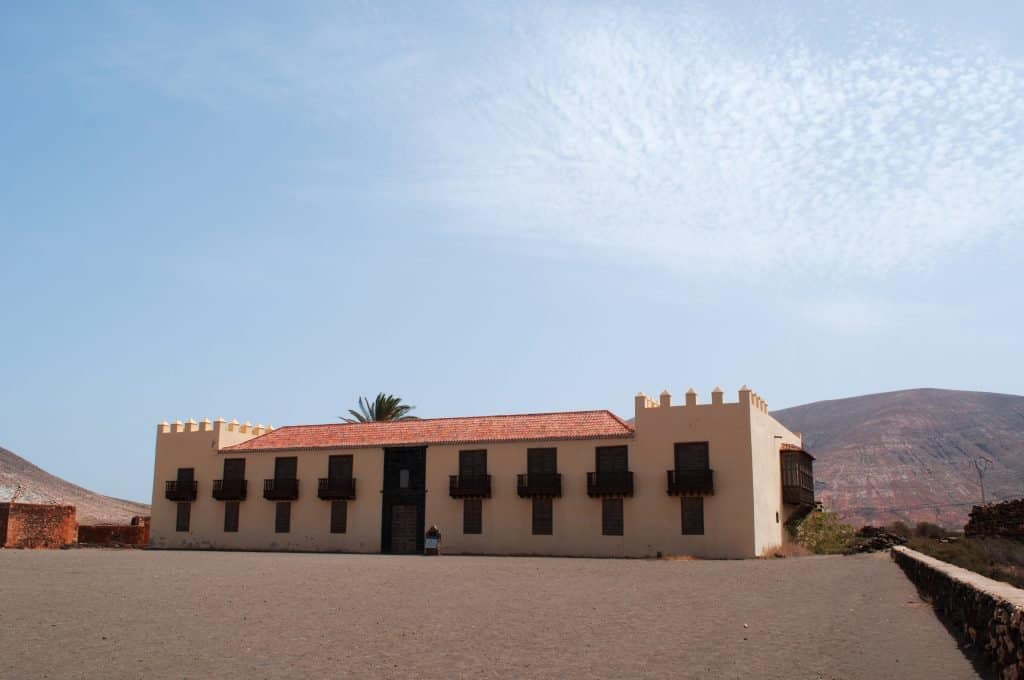 A beige building in Fuerteventura with a red-tiled roof and black balconies stands in a barren landscape under a partly cloudy sky, with hills in the background.