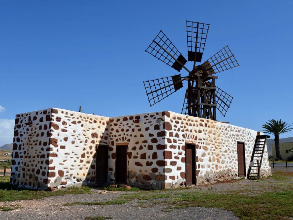A traditional windmill in Fuerteventura with a stone base and wooden blades stands in a rural area under a clear blue sky.