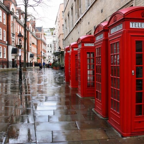 A row of London red telephone booths lines a wet, empty street with historic brick buildings on either side.