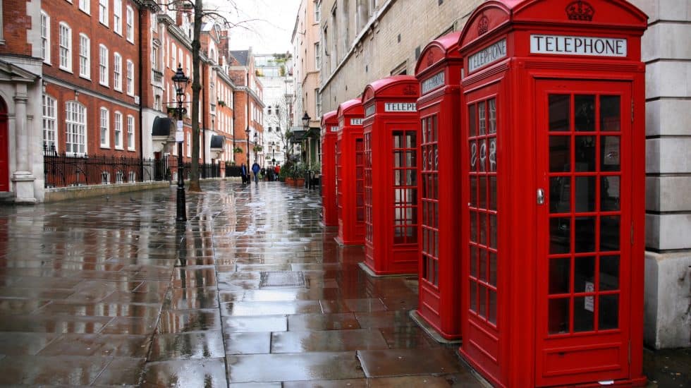 A row of London red telephone booths lines a wet, empty street with historic brick buildings on either side.