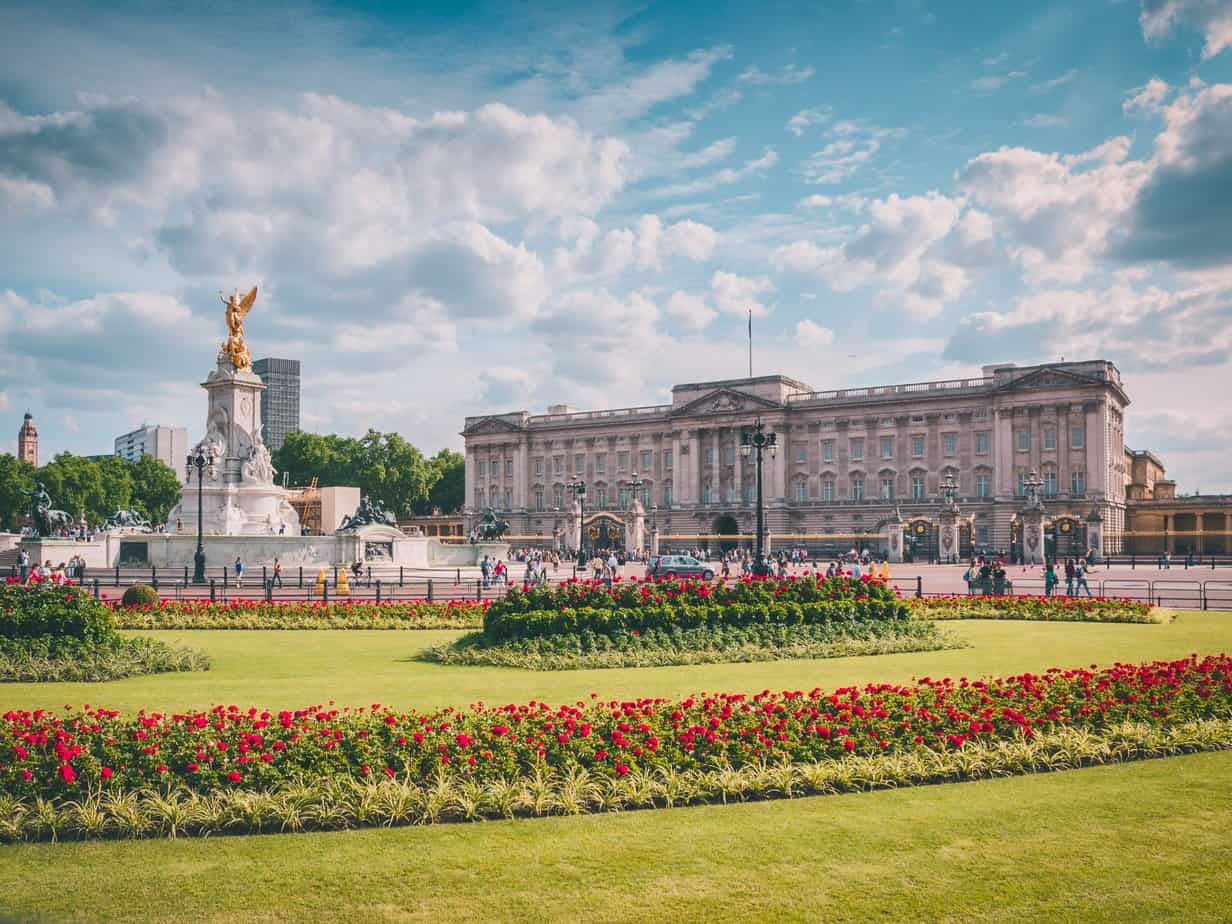 Buckingham Palace with the Victoria Memorial in front, viewed from the garden with well-maintained flower beds, under a partly cloudy sky.