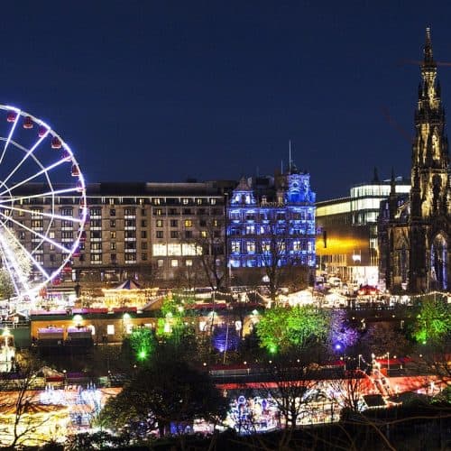 A nighttime cityscape featuring a lit Ferris wheel, colorful lights, historic buildings, and a tall, illuminated monument. Trees and other festive lights add to the vibrant atmosphere.