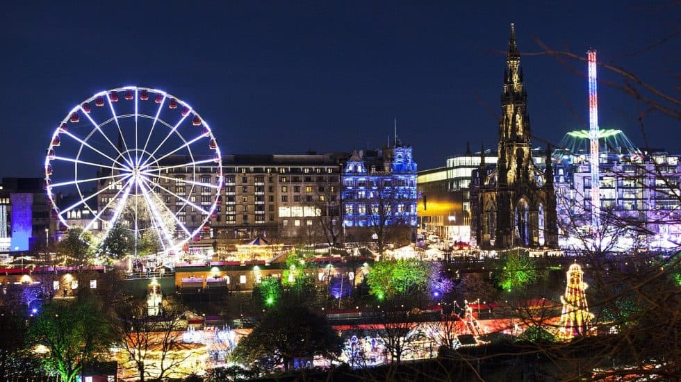 A nighttime cityscape featuring a lit Ferris wheel, colorful lights, historic buildings, and a tall, illuminated monument. Trees and other festive lights add to the vibrant atmosphere.