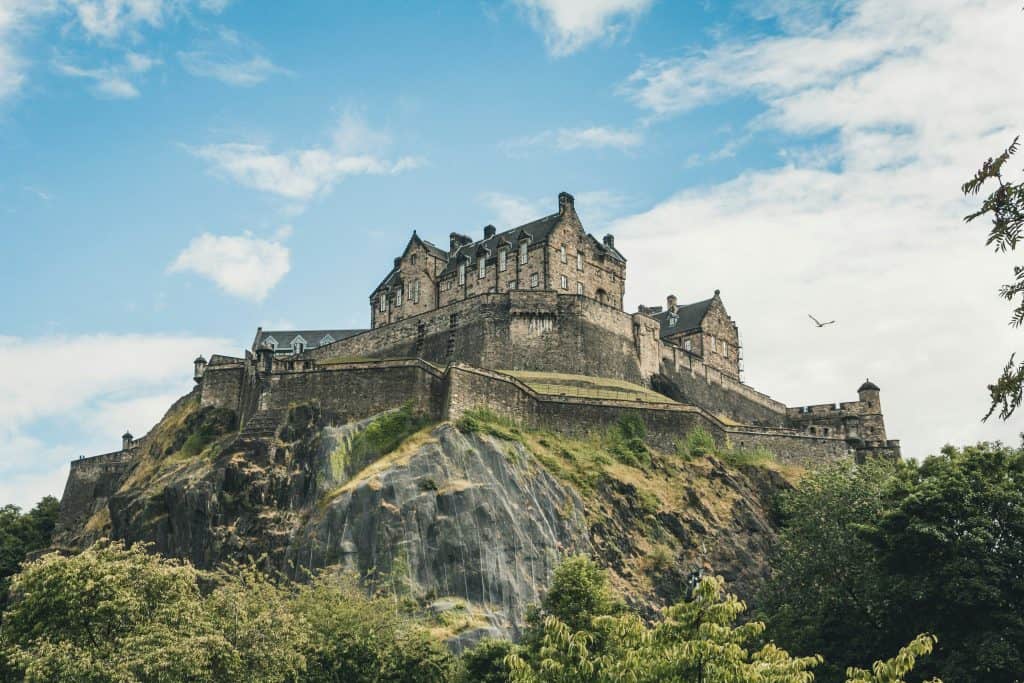 Large stone castle situated atop a rocky hill, surrounded by greenery, under a partly cloudy sky with a single bird flying.