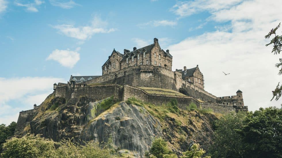 Large stone castle situated atop a rocky hill, surrounded by greenery, under a partly cloudy sky with a single bird flying.