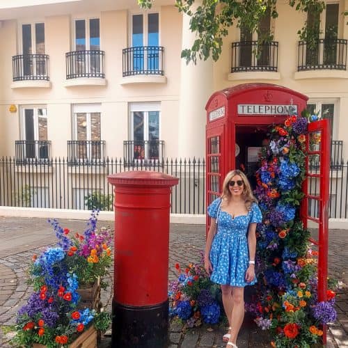 A woman in a blue dress stands next to a red telephone booth in London filled with colorful flowers beside a red post box. Flowers also spill from wooden crates on the ground.