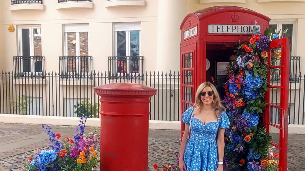 A woman in a blue dress stands next to a red telephone booth in London filled with colorful flowers beside a red post box. Flowers also spill from wooden crates on the ground.