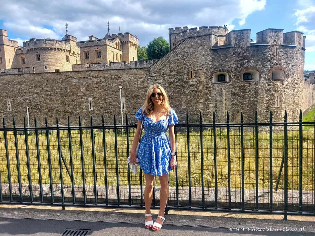 A woman in a blue dress stands in front of a historic stone castle in London with a black metal fence in the foreground. The sky above is partly cloudy.