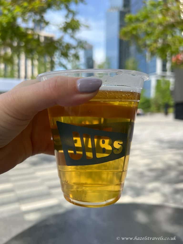 A hand holding a plastic cup of beer with "JIBs" printed on it, outdoors with buildings and trees in the background in Hudson Yards NYC.