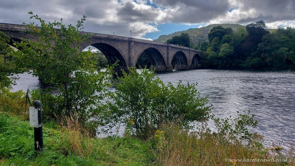 A stone bridge with multiple arches spans a wide river at Dunkeld surrounded by lush greenery and trees under a cloudy sky.